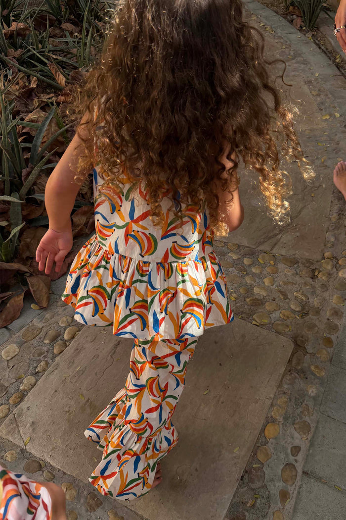 Cream cotton co-ord with a multicoloured banana print, peplum top and flared trousers on a young girl walking.