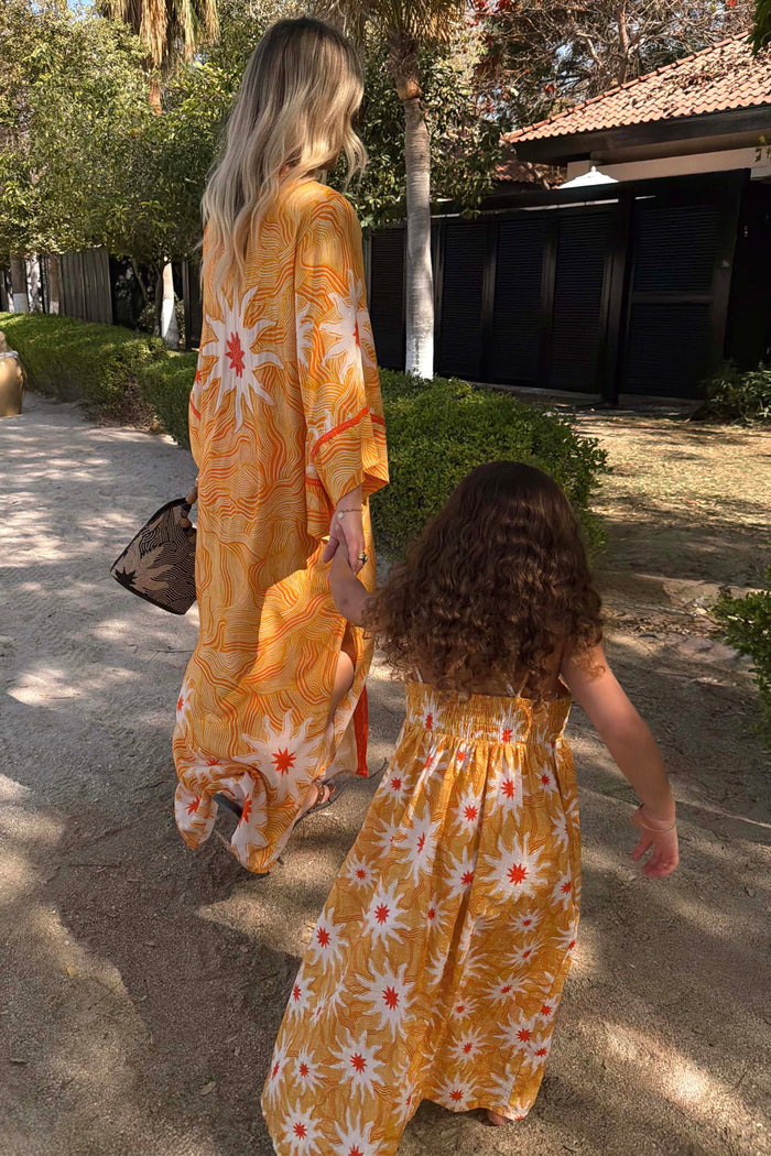 Yellow Cotton Sun Dress with an orange sun print and shirred top, on a girl in a sunny vacation setting.