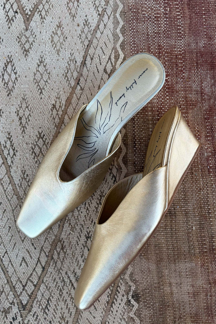 Metallic gold leather Wedge Mules with a pointed toe and sculpted heel, designed in London and shown on a rug.