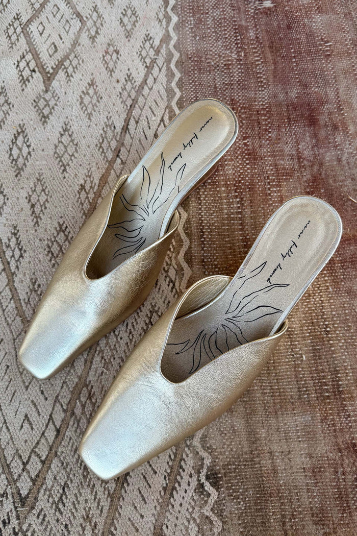 Gold metallic leather wedge mules with a pointed toe and open back, resting on a textured rug.