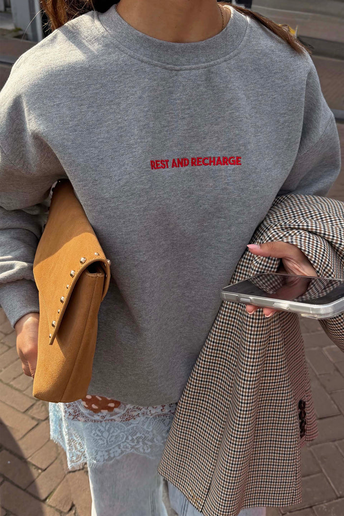 Grey Rest and Recharge Jumper with red embroidered text, styled with a tan suede bag and a checked blazer.