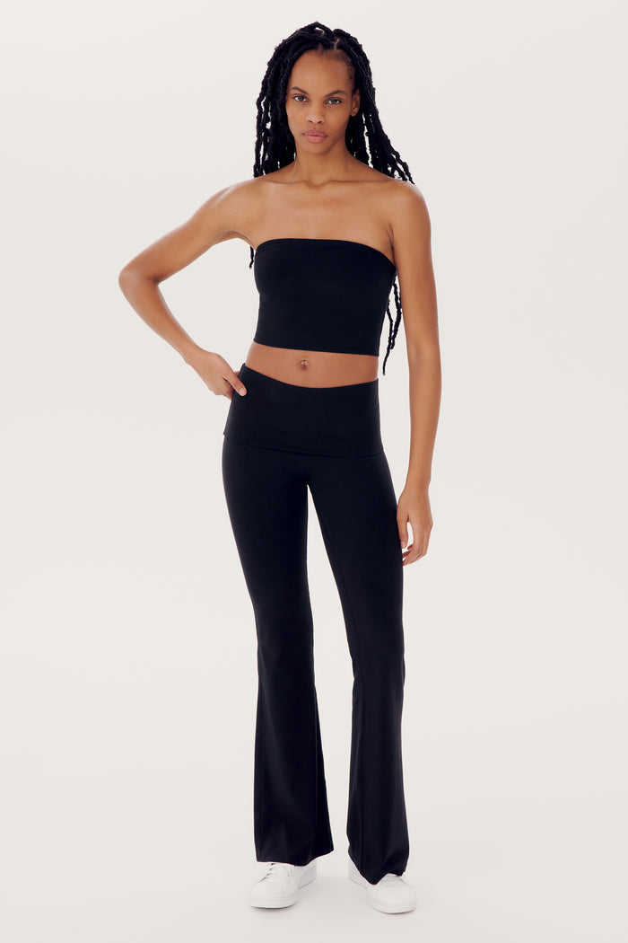 A person with long hair in a long braid stands against a plain background wearing the SPLITS59 Hazel Airweight Crop Bandeau - Black, black flared spandex pants, and white shoes.
