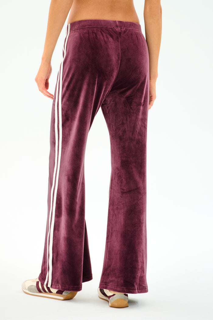 A model wears SPLITS59 Heritage Velour Pant in Dark Cherry/White, made from a soft polyester-spandex blend, with white side stripes and beige sneakers, shown from the back against a plain white background.
