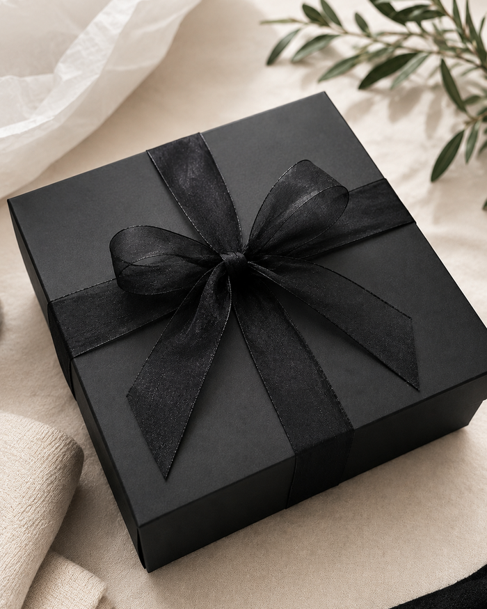 A square black gift box wrapped with a wide black ribbon and bow, styled on a light beige fabric surface with soft white tissue paper and green leaves in the background.