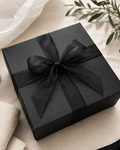 A square black gift box wrapped with a wide black ribbon and bow, styled on a light beige fabric surface with soft white tissue paper and green leaves in the background.