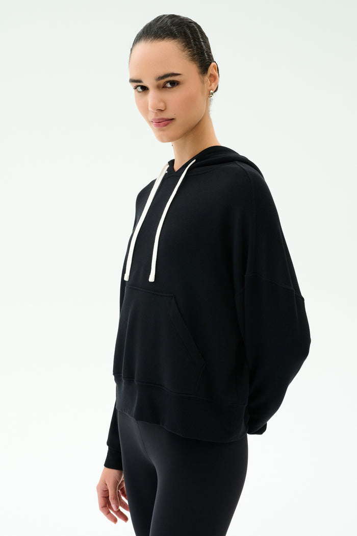 Person wearing the SPLITS59 Joey Fleece Hoodie in black—crafted from comfortable modal fabric for a soft, elevated feel—with white drawstrings and black pants, standing against a plain white background.