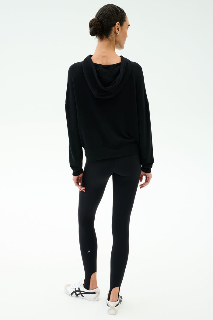 A woman stands with her back to the camera, wearing the SPLITS59 Joey Fleece Hoodie in black and matching leggings, paired with white sneakers, against a plain white background.