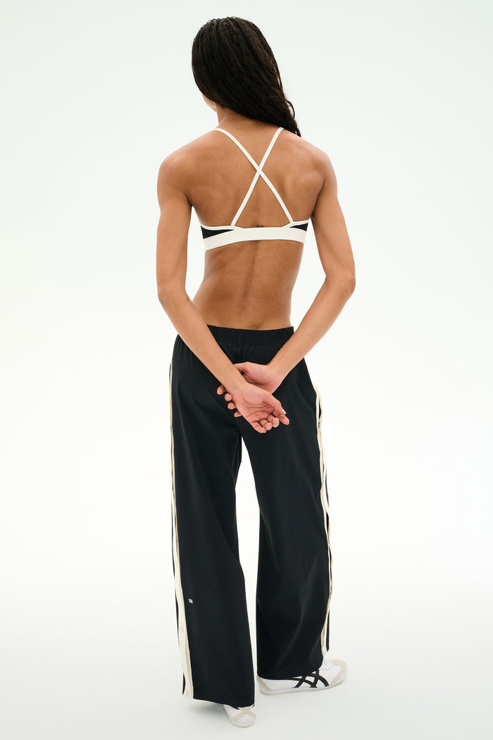 A person stands with hands behind their back, facing away from the camera against a plain background, wearing black SPLITS59 Jordan Nylon Twill Tearaway pants in Black/Creme featuring white stripes, paired with a white strappy sports bra and white sneakers.