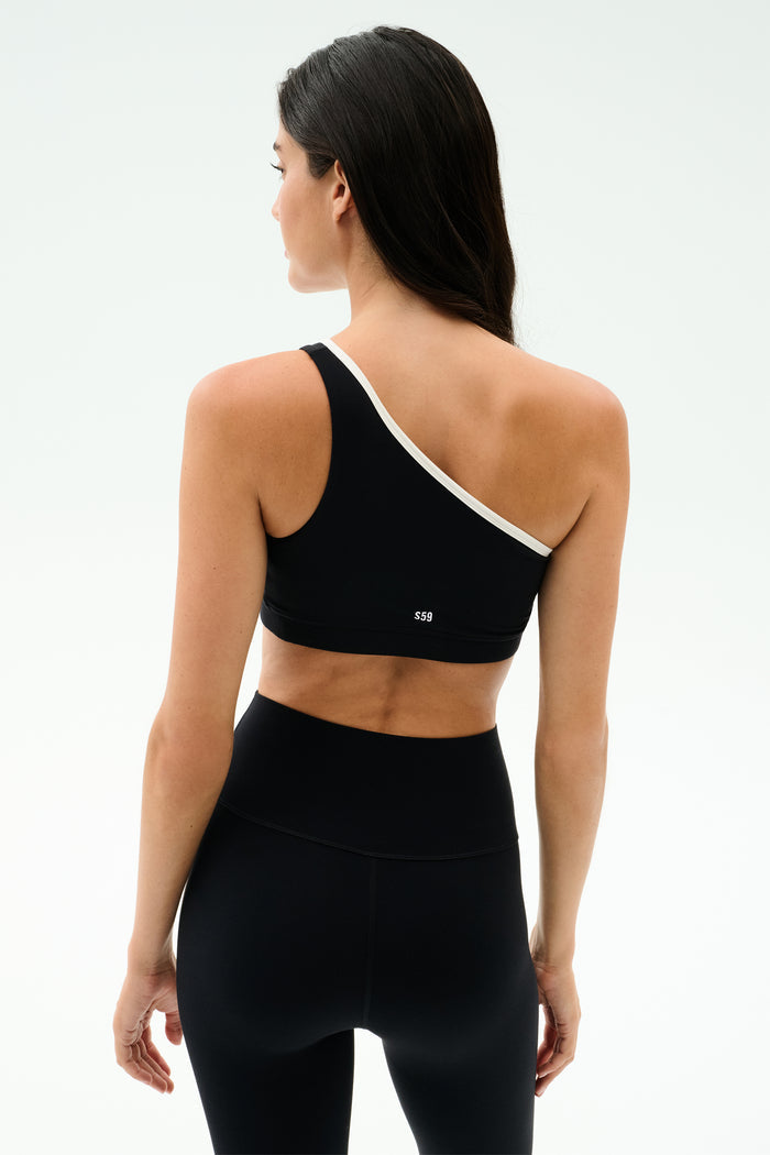 A woman faces away, wearing the SPLITS59 Josie Airweight One Shoulder Bra in Black/White with matching high-waisted leggings, set against a plain light background.