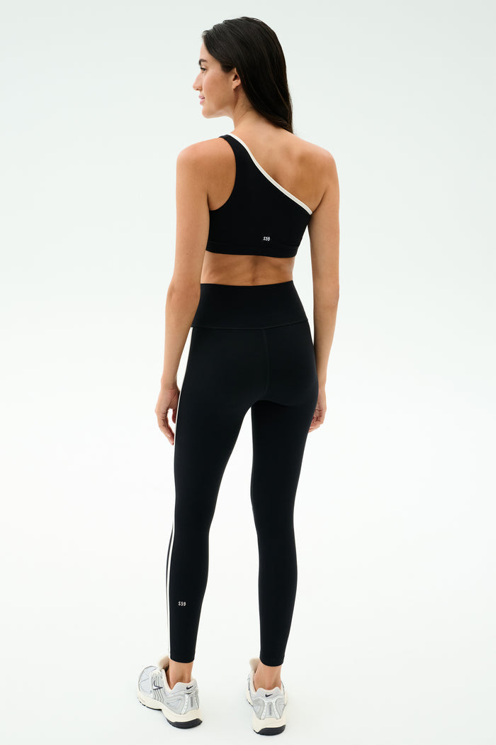 A woman stands facing away on a light background, wearing the SPLITS59 Josie Airweight One Shoulder Bra in black/white with matching black leggings featuring white side stripes, paired with white athletic shoes.