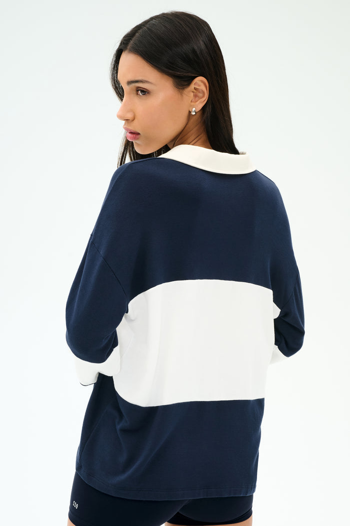 A woman with long dark hair models the SPLITS59 Katie Fleece Rugby in Indigo/White, glancing over her shoulder against a plain light background.