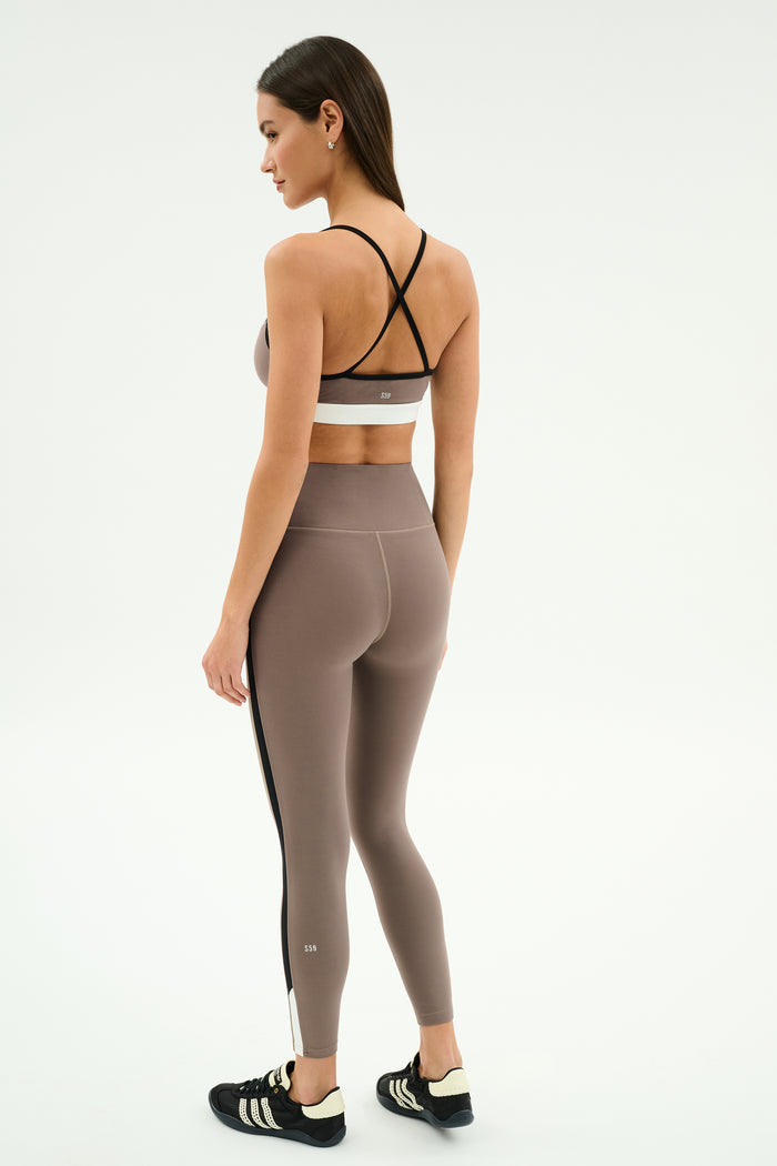 A woman stands facing away, wearing the SPLITS59 Layla Airweight High Waist 7/8 leggings in Lentil/Black, featuring black crisscross straps and ultra-light Airweight fabric—ideal for hot yoga—paired with black athletic shoes.