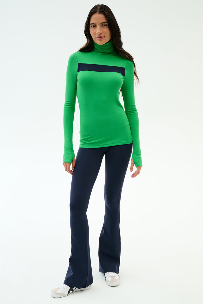 A woman faces forward wearing the SPLITS59 Lennon Rib Turtleneck in Grass/Indigo and navy flared pants with white sneakers—ideal for yoga or errands—against a plain white background.