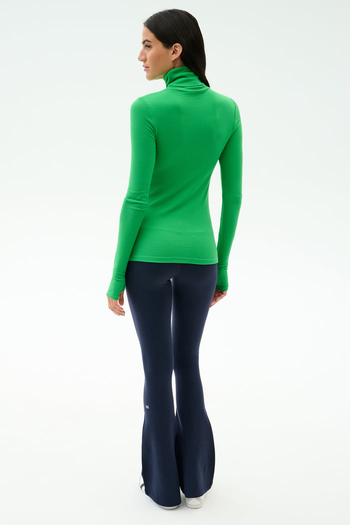 A person stands facing away, wearing the SPLITS59 Lennon Rib Turtleneck in Grass/Indigo and dark flared pants against a plain, light background.