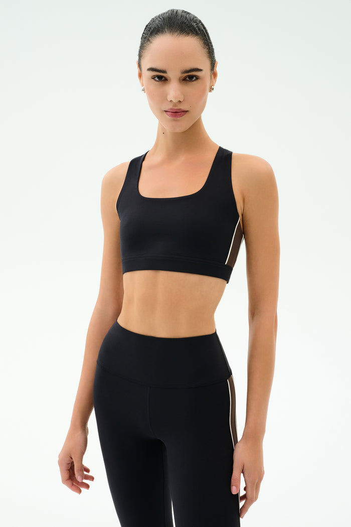 A woman stands facing forward in the SPLITS59 Lexi Rigor Bra - Black/Dark Chocolate, ideal for gym workouts, against a plain white background.