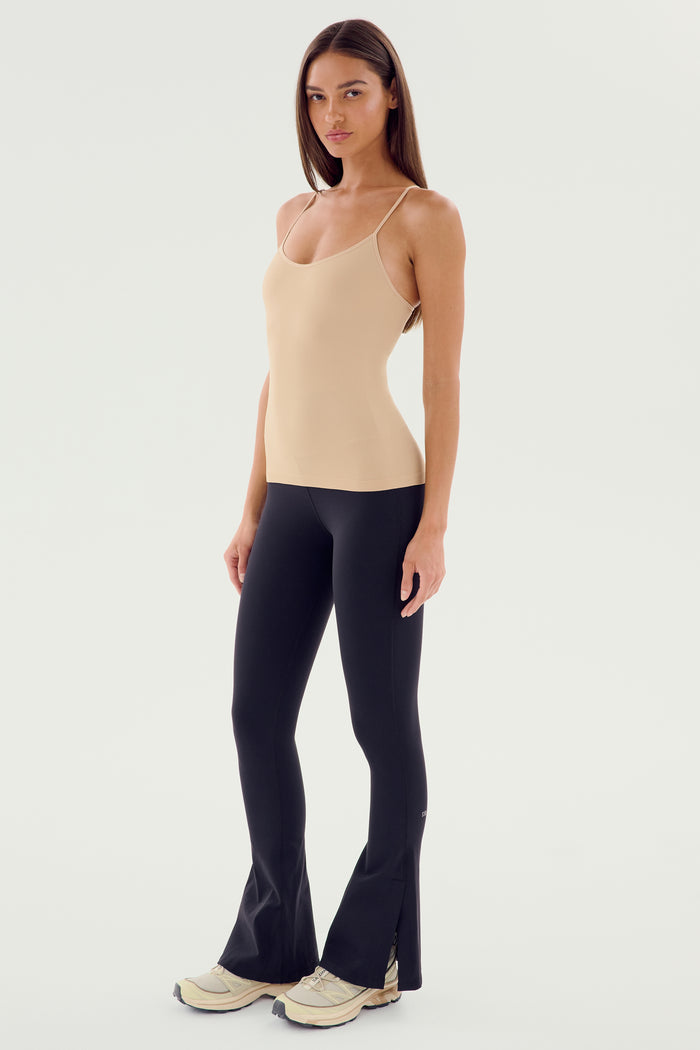 A woman dressed in the SPLITS59 Loren Seamless Waist Length Tank - Nude and black leggings stands against a plain white background. She has long hair, poses with one foot slightly forward, and is wearing white shoes, ready for her gym workouts.