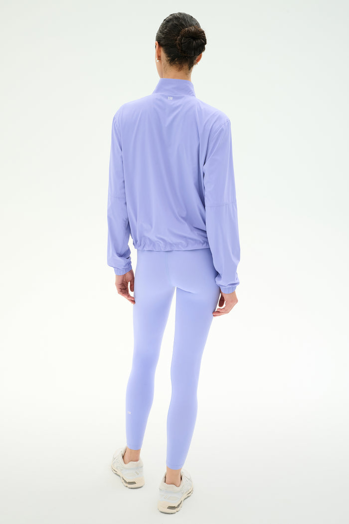 A person stands with their back to the camera, wearing the SPLITS59 Lyle Woven Nylon Pullover in Purple Haze/Creme and matching leggings with white sneakers against a plain white background.
