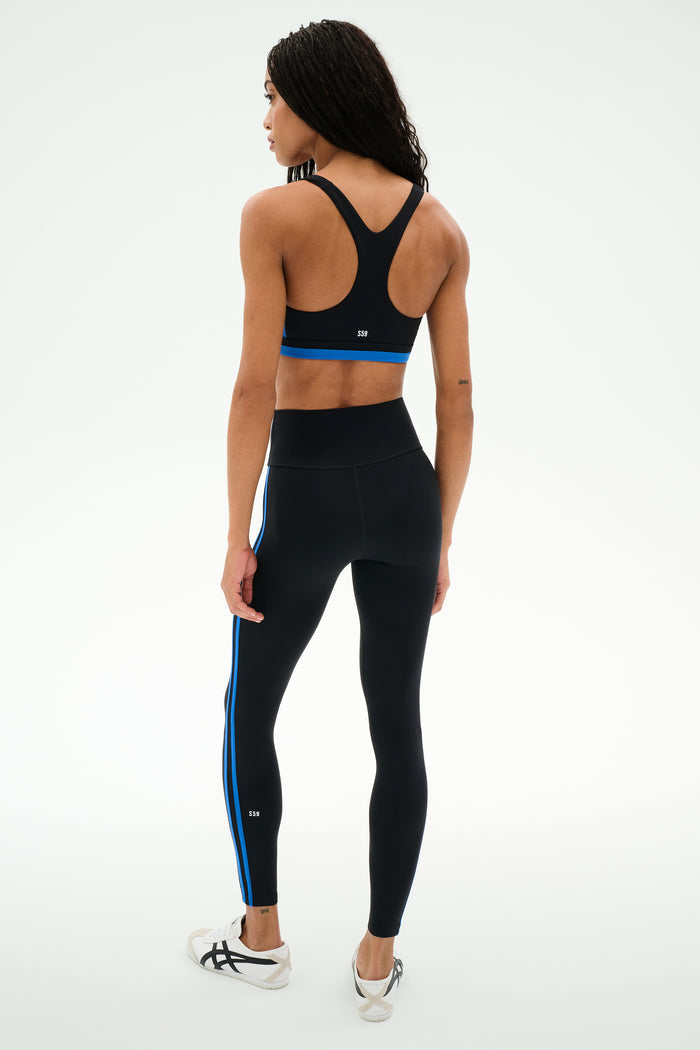 A person stands facing away against a plain white background, wearing the SPLITS59 Maci Airweight Bra in Black/Classic Blue.