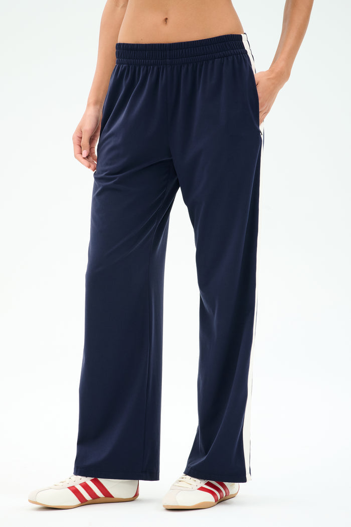A person wearing SPLITS59 Maxie Rigor Track Pant in Indigo/White with white sneakers featuring red stripes stands against a plain background.