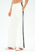 A person wears SPLITS59 Maxie Rigor Track Pant in White/Indigo, featuring a navy side stripe, paired with white sneakers with red stripes, standing against a plain background.