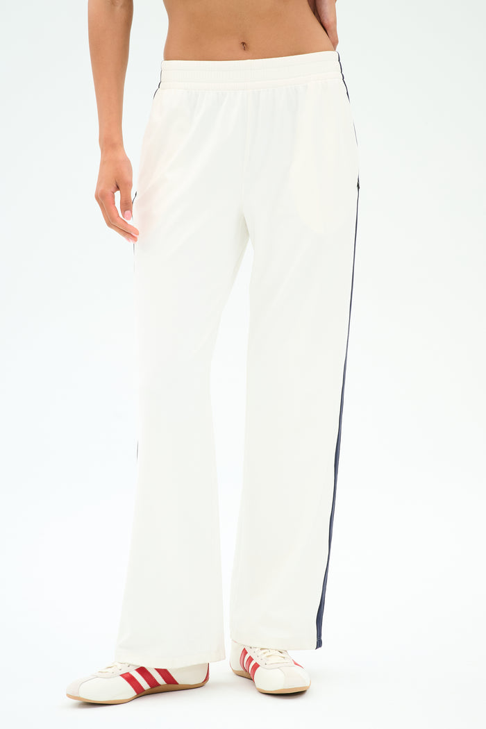 A person wears the SPLITS59 Maxie Rigor Track Pant in White/Indigo with black side stripes and white sneakers with red stripes, standing against a plain light background. Made from soft Rigor fabric for comfort and style.