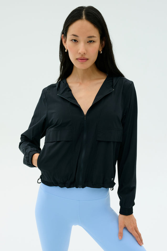A woman faces forward against a plain light background, wearing the SPLITS59 Megan Woven Nylon Jacket in black and light blue spandex leggings.