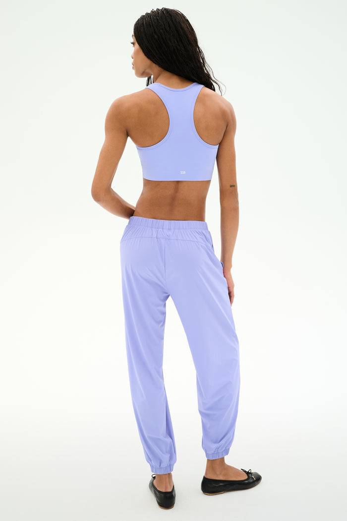 A person with long braided hair faces away from the camera, wearing SPLITS59’s Megan Woven Nylon Jogger in Purple Haze and a matching lavender sports bra, styled with black flats against a plain white background.