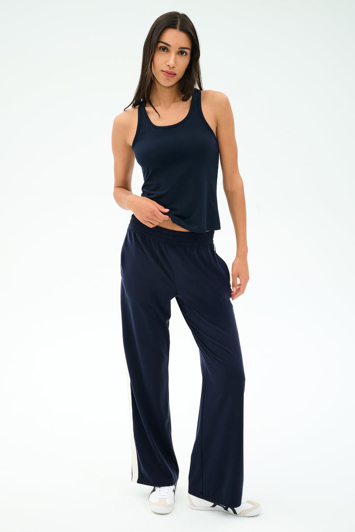 A woman stands against a plain background wearing the SPLITS59 Mel Airweight Lite Tank in Indigo, matching wide-leg pants with a white stripe, and white sneakers.