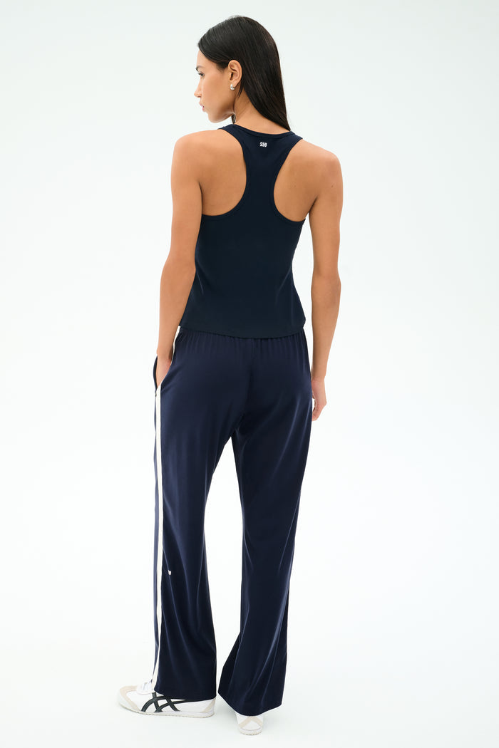 A woman faces away, wearing the SPLITS59 Mel Airweight Lite Tank in Indigo with matching wide-leg pants, both featuring white side stripes, paired with white sneakers.