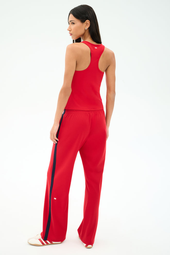 A woman faces away, wearing the SPLITS59 Mel Airweight Lite Tank in Pirate Red with matching track pants featuring a navy side stripe, paired with white sneakers.