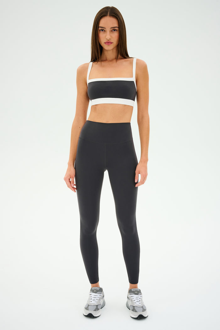 A person stands in a gray and white athletic outfit, ready for gym workouts. The sleek design of the SPLITS59 Monah Rigor Bra - Graphite/White adds style against the plain background.