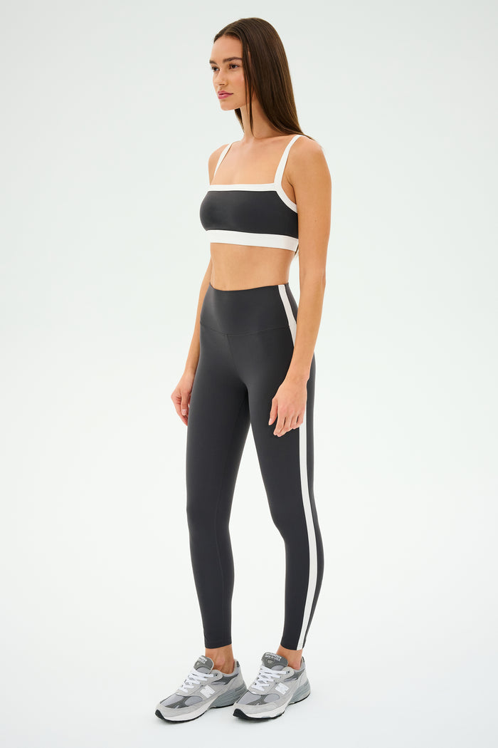 A woman confidently poses in a SPLITS59 Monah Rigor Bra in graphite/white and silver sneakers against a plain background, capturing the essence of gym workouts.