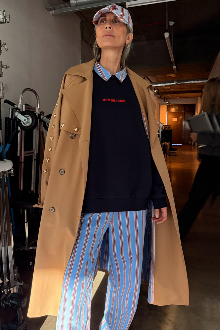 Navy Book The Flight Jumper with red text, styled with a tan trench coat and blue striped trousers for a London look.