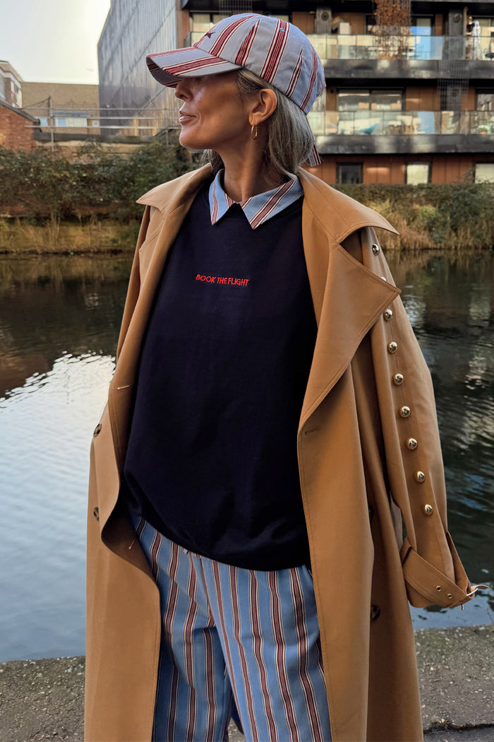 Navy Book The Flight Jumper with red slogan text, layered under a tan trench coat with striped trousers.