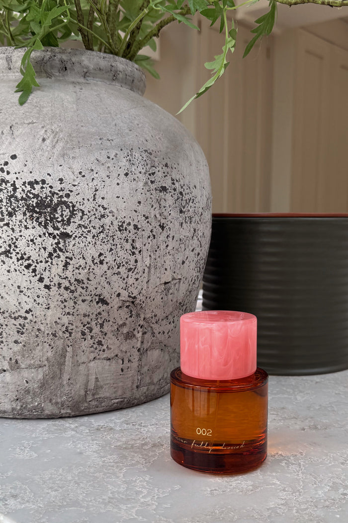 NFD Eau De Parfum 002 in an amber glass bottle with a pink marbled lid, styled beside a rustic grey stone vase.