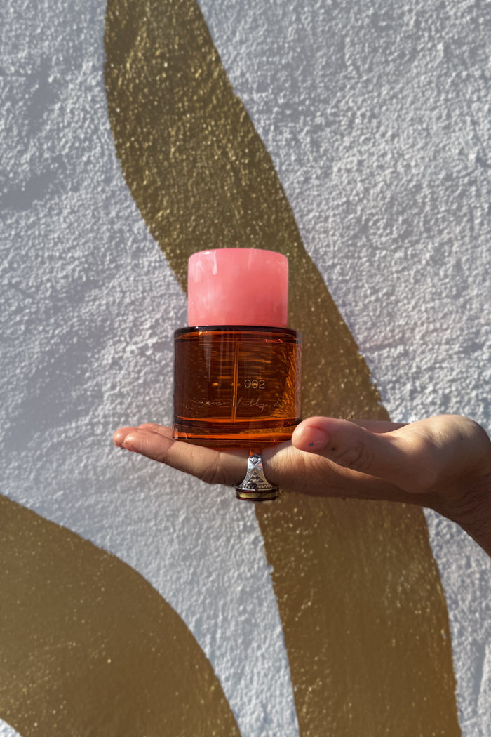 NFD Eau De Parfum 002 amber glass bottle with a pink marbled cap, held in a palm against a textured gold wall.