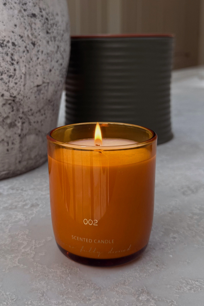 Lit NFD Scent 002 Candle in a warm orange glass jar with gold branding, set on a textured marble-style surface.