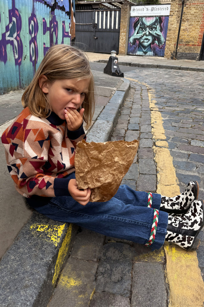 NFD Kids Aspen Jumper with a geometric knit pattern, worn with jeans and cow-print boots on a cobbled London street.