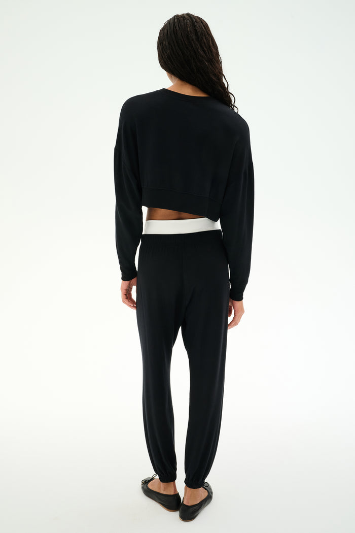 A person with long hair faces away, wearing the SPLITS59 Noah Fleece Crop Sweatshirt in black, paired with black pants and flats, set against a plain white background.