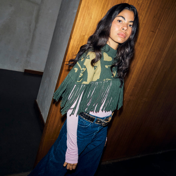 Olive Amara Knit Cape with a green heart print and tassel fringe, styled with a pink top and denim jeans.