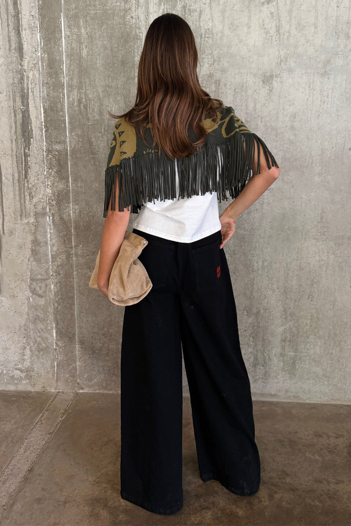 Olive Amara Knit Cape with tassel fringe and printed motifs, seen from the back with black wide-leg trousers.