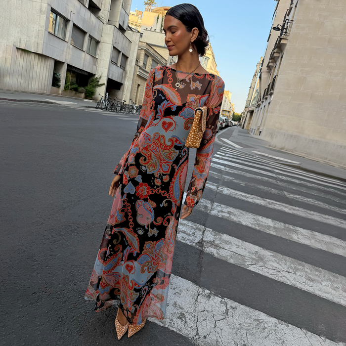 A woman stands on a city street crosswalk wearing a long-sleeve maxi dress with a colorful paisley and floral print in pink, orange, blue, and black. She is holding a small brown woven handbag and wearing pointed-toe heels.