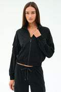 A woman with long brown hair wears the SPLITS59 Peaches Airweight Cargo Jacket in black, paired with matching black stretch pants, standing against a plain white background.