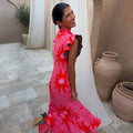 Pink Soleil Remi Dress with red sun motifs and leopard-lined pleated frills on a smiling woman in a courtyard.