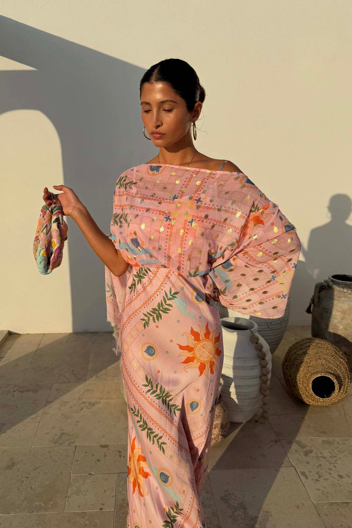 Pink Sun Anais Dress featuring a sheer draped top, gold sun motifs and a bias cut skirt on a sunny outdoor patio.