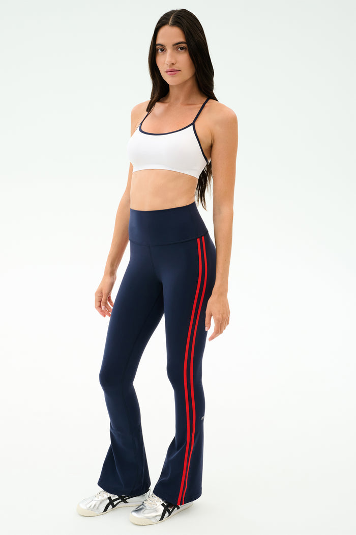 A woman wears the SPLITS59 Raquel High Waist Flared Legging in Indigo/Lotus Red, paired with a white sports bra and white sneakers, standing against a plain light background. The leggings offer 4-way stretch for comfort and performance.
