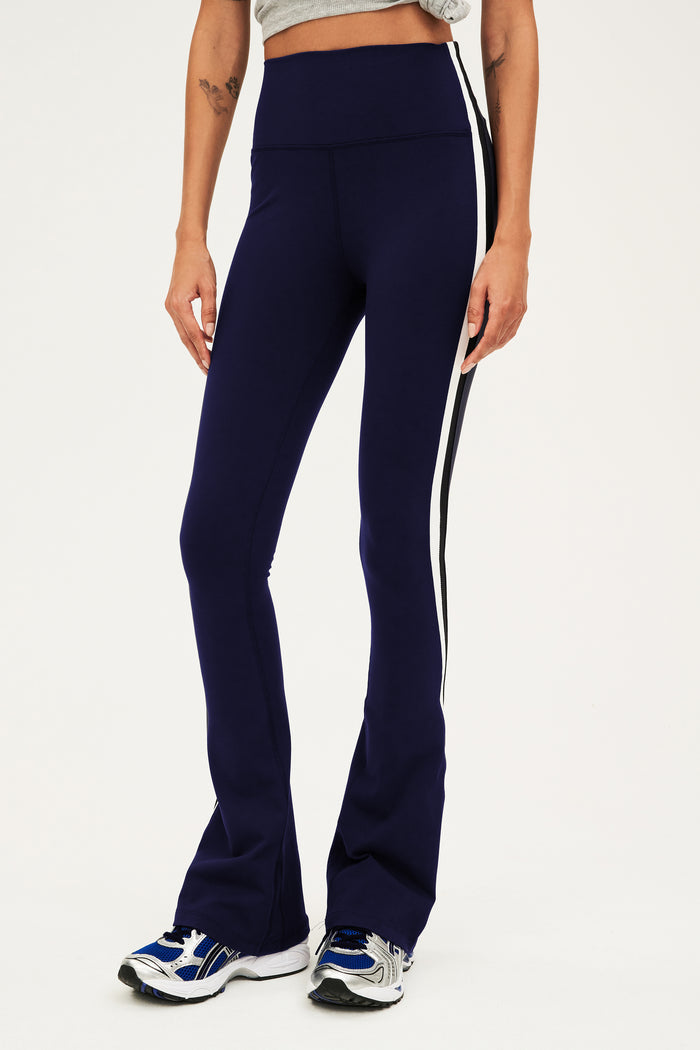 Front view of woman wearing dark blue high waist below ankle length legging with wide flared bottoms and white and black side  stripes on both legs. Paired with gray,black and bright blue shoes.