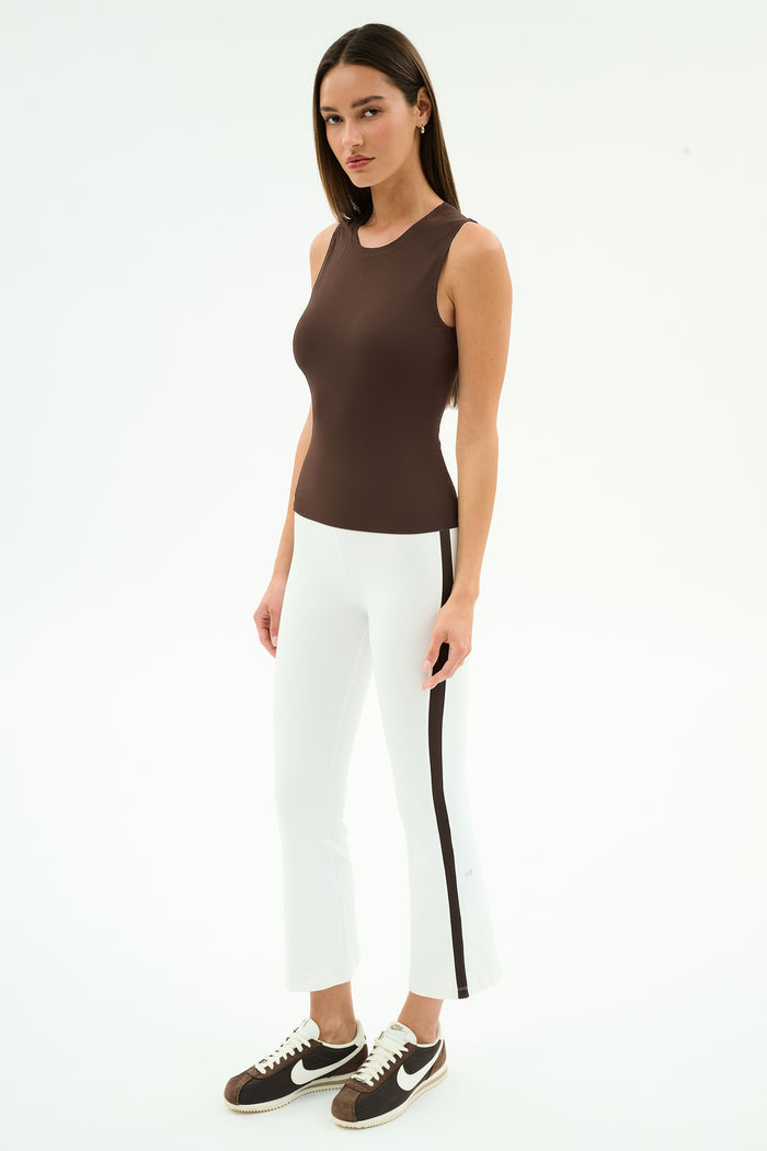 A woman wearing a sleeveless brown top, SPLITS59 Raquel High Waist Supplex Crop leggings in White/Dark Chocolate, and brown sneakers stands against a plain white background.