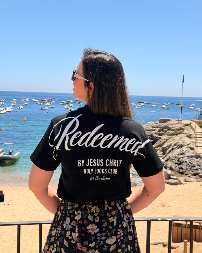 A woman stands on a beach facing the ocean, wearing a black t-shirt with 'Redeemed BY JESUS CHRIST HOLY LOOKS CLUB ft. the chums' printed in white on the back, paired with a black floral skirt.