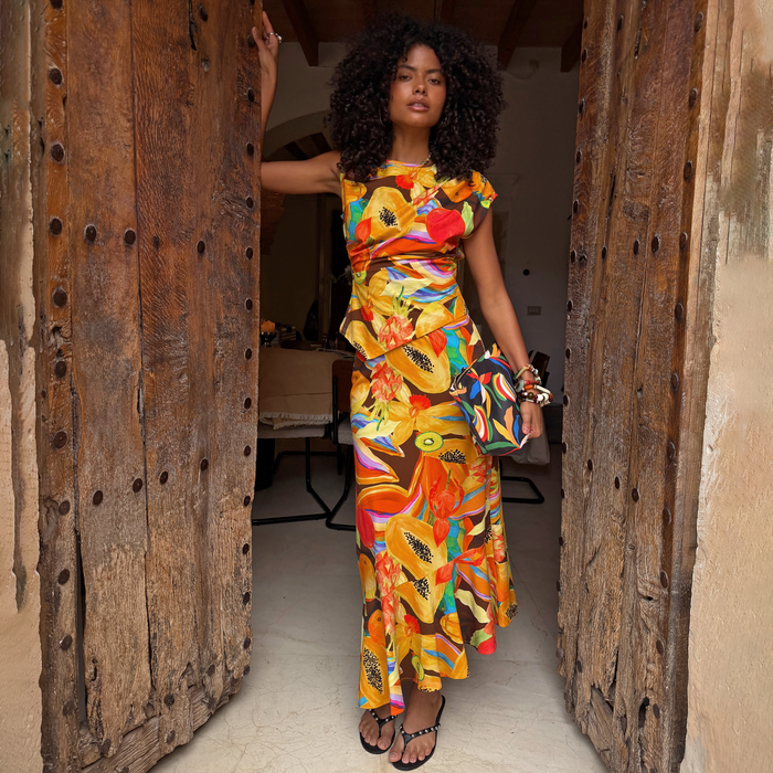 Rio A-Symmetric Skirt in tropical fruit print, styled as a vacation co-ord in a rustic doorway.